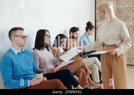 HR manager with blond hair giving clipboard with questionnaire to job applicants sitting on chairs against wall in corridor Stock Photo