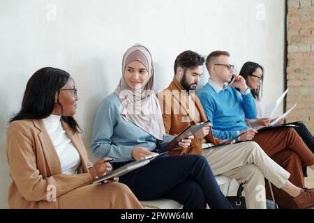 Smiling young Black and Arabian women sitting on chairs in corridor with other job candidates and discussing vacancy while being in line Stock Photo