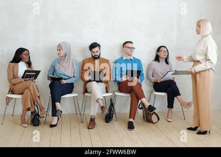 Blond-haired recruitment manager standing in front of interracial job applicants sitting in row and explaining job interview process Stock Photo