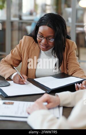 Smiling young Afro-American woman sitting at table and signing contract while accepting job offer after interview Stock Photo