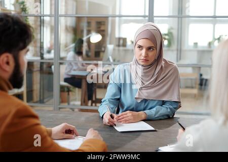 Serious young Arabian woman in hijab sitting at table in front of HR managers and filling questionnaire at job interview Stock Photo