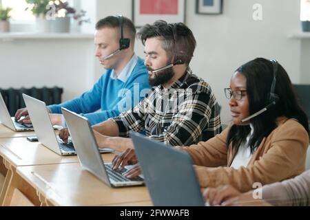 Group of concentrated young multi-ethnic call center agents sitting at tables and using laptops while selling products by phones Stock Photo