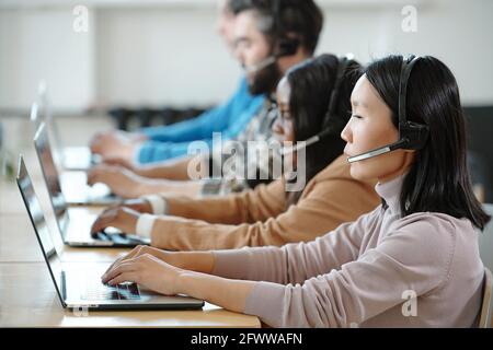 Focused young interracial operators in headsets with microphone sitting in row and answering incoming calls in contact center Stock Photo