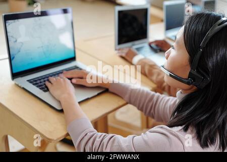 Over shoulder view of busy young Asian call center operator in wireless headset with microphone providing customer with information using laptop Stock Photo