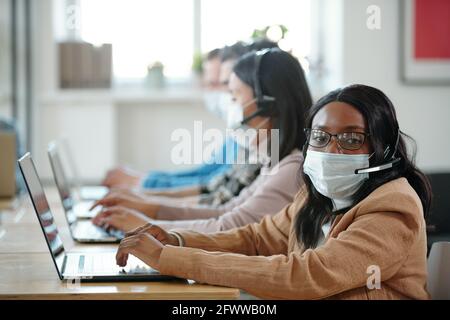 Portrait of young African-American female technical support specialist in mask and eyeglasses sitting at table with laptop in contact center Stock Photo