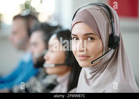 Portrait of smiling beautiful Arabian contact center agent in hijab using wireless headset with microphone while answering customers Stock Photo