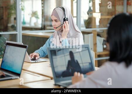 Serious young Arabian call center agent adjusting headset microphone while responding to customer using laptop in office Stock Photo