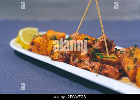 Aperitif or typical Spanish tapa, baby squid with garlic and potatoes, served on an elongated white tray with lemon and rustic background. Traditional Stock Photo