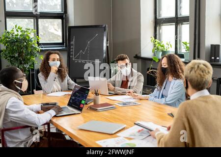 Group of colleagues in protective masks discussing working points at meeting in office Stock Photo