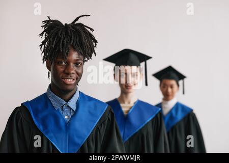 Diverse group of three happy college graduates wearing robes and smiling at camera while standing in row Stock Photo