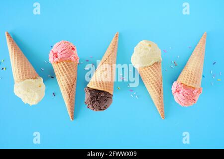 Ice cream cone flat lay over a blue background. White vanilla, pink strawberry and dark chocolate flavors. Stock Photo