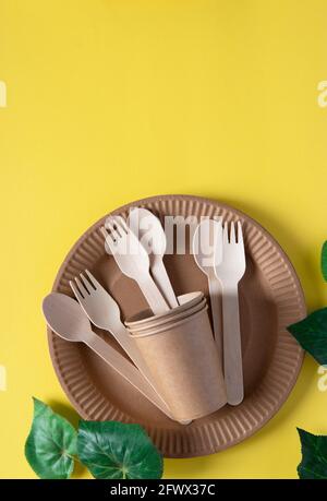 Wooden forks and spoons in a paper cup with paper plate on a yellow background. Zero waste concept. Vertical format. Top view Stock Photo