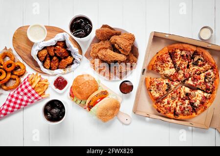 Table scene with large variety of take out and fast foods. Hamburgers, pizza, fried chicken and sides. Top view on a white wood background. Stock Photo