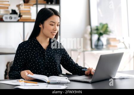 Successful beautiful smart young asian woman, freelancer or student sitting at the desk, using laptop for work or online study, taking notes during webinar or online lecture, smiling friendly