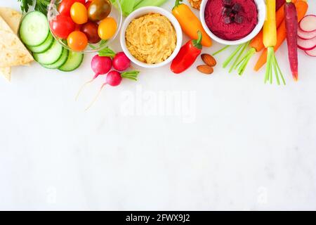 Assortment of fresh vegetables and hummus. Overhead view top border on a white marble background. Stock Photo