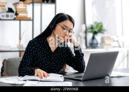 Upset young beautiful asian woman in glasses and headset, call center worker, manager or freelancer, sitting at table in the office, looking sadly at the laptop screen, resting her head on her hand Stock Photo