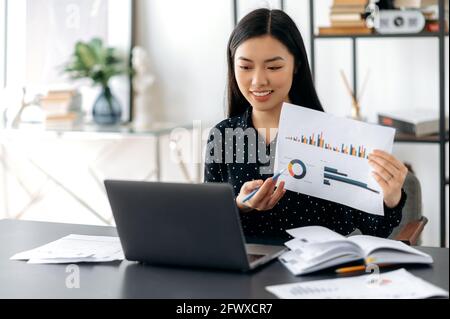 Friendly confident asian business woman or coach in formal shirt, sitting at her desk, analyzing report, showing financial graph to employees during video communication over conference call, smiling Stock Photo