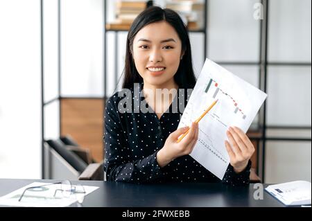 Successful attractive smart confident asian business woman, financial coach or top manager sitting at table, talks with colleagues or clients by video link, showing charts, conducts consultation,smile Stock Photo