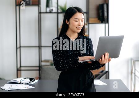 Portrait attractive confident successful smart pretty young asian business lady or manager standing near work desk in office, in formal clothes, holds a laptop in hands, browsing news, smiles Stock Photo