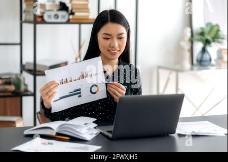 Young adult friendly smiling confident asian woman in formal shirt, sitting at her work desk, analyzing report, showing financial graph to colleagues during communicating by video conference using Stock Photo