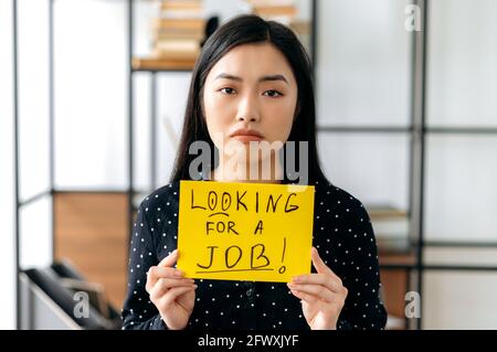 Portrait depressed unemployed young Asian woman, student or freelancer, sits at table with sign with inscription looking for a job, hopes to get job of her dreams, look sadly at camera Stock Photo