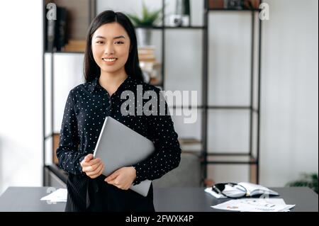 Portrait confident successful smart joyful pretty young asian woman. Female japanese girl, office manager, stand near work desk in office, holds laptop in hands, looks at camera, smiles friendly Stock Photo