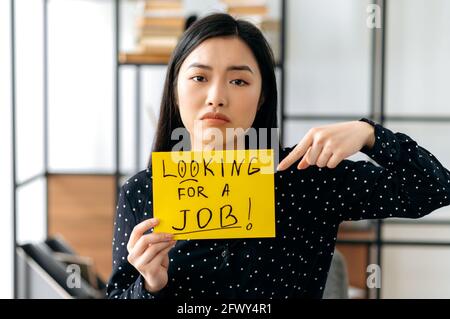 Upset unemployed asian woman student or freelancer, sits with sign with inscription looking for a job, hoping to get a dreams job, look sadly at camera, pointing her finger at inscription Stock Photo