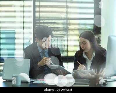 View through glass wall of two businesspeople talking to each other while working together in office room Stock Photo