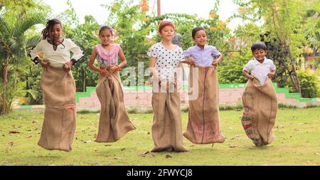 Group of Childrens playing potato sack jumping race at park outdoor during summer camp - kids having fun while playing gunny sack racing competition. Stock Photo