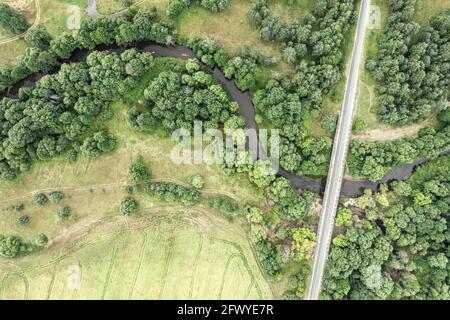 country road with bridge over winding river flowing through green forest and cultivated fields. aerial top down view
