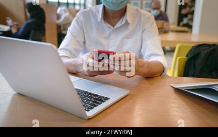 Man with face mask reading phone with computer laptop on table in a library. Close up shot. Stock Photo