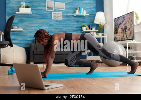 Black fit athletic woman training for muscle strenght doing mountain climbers position on yoga mat dressed in sportwear leggings, in home living room following online instructions. Stock Photo