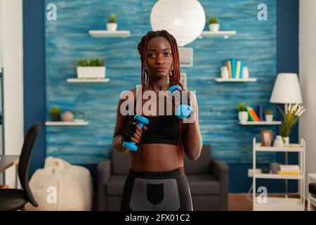 Athletic fit strong black woman working out with with dumbbells standing in home living room for healthy lifestyle, taking care of body exercising, using modern equipment. Stock Photo