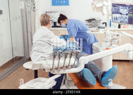 Stomatologist senior woman doing hygienic cleaning of teeth using sterile dental tools. Patient sitting with open mouth on dental chair, dentist doctor performing examination in modern clinic Stock Photo