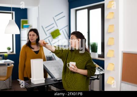 Team of women architects standing in creative agency office discussing and analysing building model measuring prototype with pencil. Engineers sharing ideas for build design in multimedia workplace Stock Photo