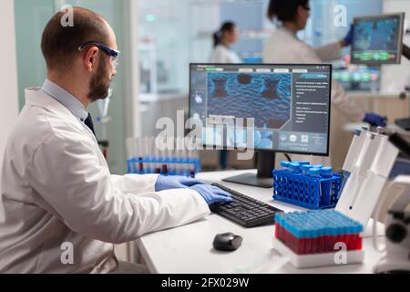 Man scientist working to develop a new vaccine in modern equipped laboratory, typing at computer. Group of biochemistry doctors examining virus evolution using high tech for researching treatment. Stock Photo