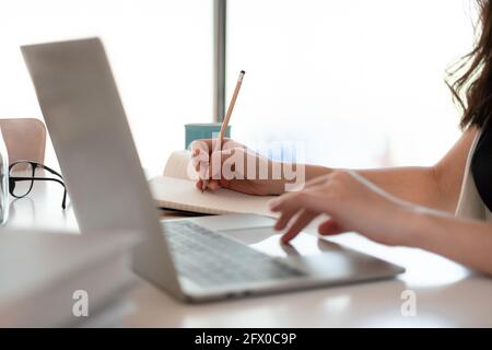 Close up woman's hands with laptop computer, notebook and pen taking notes in business office Stock Photo