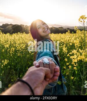 Happy young female holding boyfriend hand and looking at camera while standing on blossoming rapeseed field on sunny weather Stock Photo