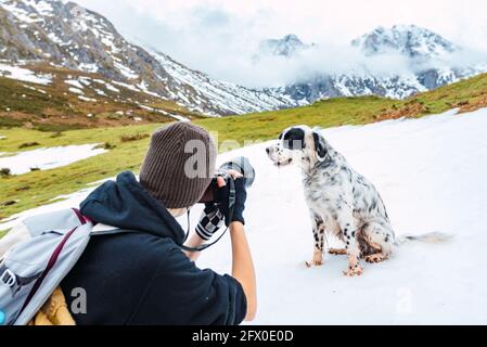 Back view of female photographer in warm clothes and with backpack using photo camera to shoot English Setter dog on snowy mountains in Peaks of Europ Stock Photo