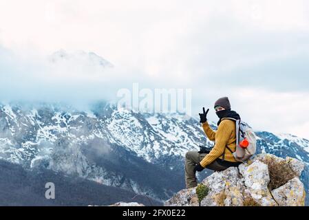 Side view of hiker with backpack showing two fingers while sitting on high point of amazing ridge of Peaks of Europe in clouds Stock Photo