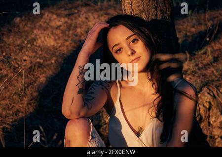 From above calm female in white dress sitting at tree trunk in dark woods in calm sundown light Stock Photo