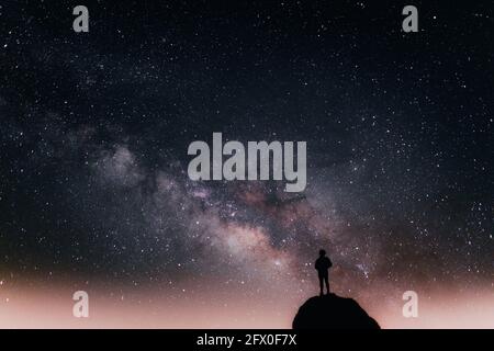 Low angle of silhouette of anonymous tourist standing on cliff against glowing starry sky at night Stock Photo