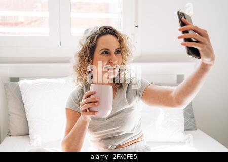 Cheerful young female with mug taking selfie on smartphone while sitting on comfy bed Stock Photo