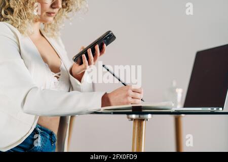 Side view of cropped unrecognizable concentrated young female entrepreneur in stylish clothes and eyeglasses using smartphone and taking notes in plan Stock Photo
