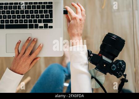 Top view of crop unrecognizable female photographer inserting memory card of photo camera into laptop sitting at glass table Stock Photo
