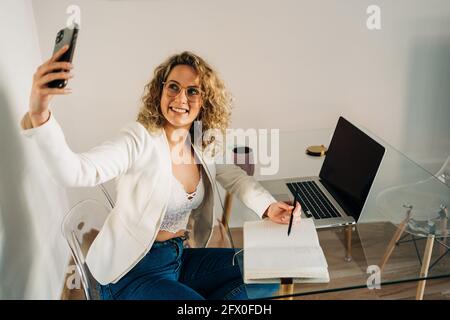 From above of cheerful young female freelancer freelancer with curly hair in stylish outfit taking selfie on smartphone while working remotely on proj Stock Photo