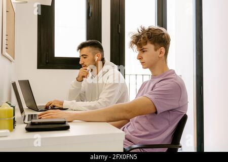 Side view of concentrated young multiethnic male colleagues in casual outfits working remotely on laptops sitting at table at home Stock Photo