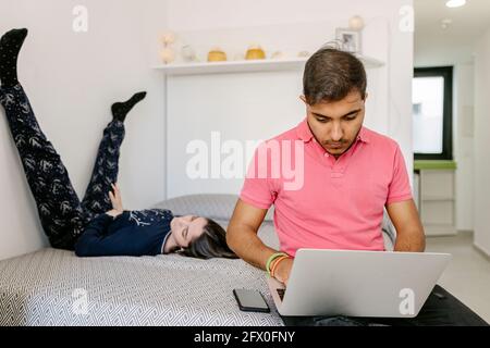 Happy young ethnic male freelancer working remotely on laptop sitting on bed near lying girlfriend browsing smartphone Stock Photo