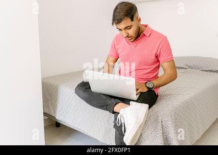 Concentrated young self employed ethnic guy in stylish outfit typing on laptop while working remotely sitting on comfortable bed at home Stock Photo