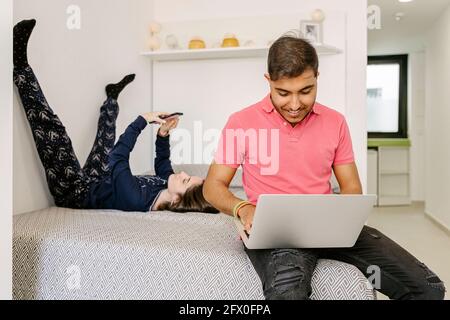 Happy young ethnic male freelancer working remotely on laptop sitting on bed near lying girlfriend browsing smartphone Stock Photo